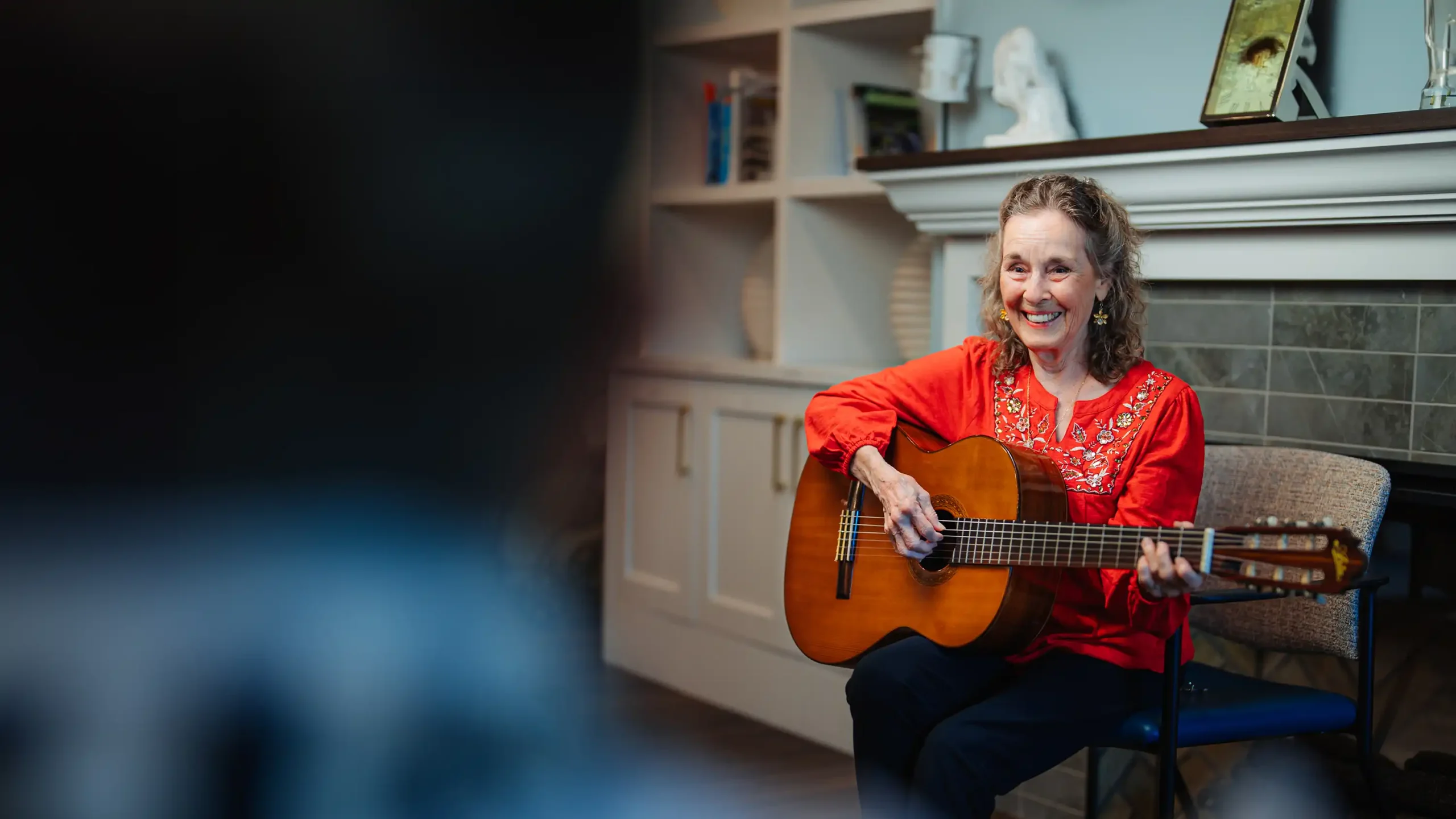 A senior woman strums a guitar by are fireplace in a living room setting