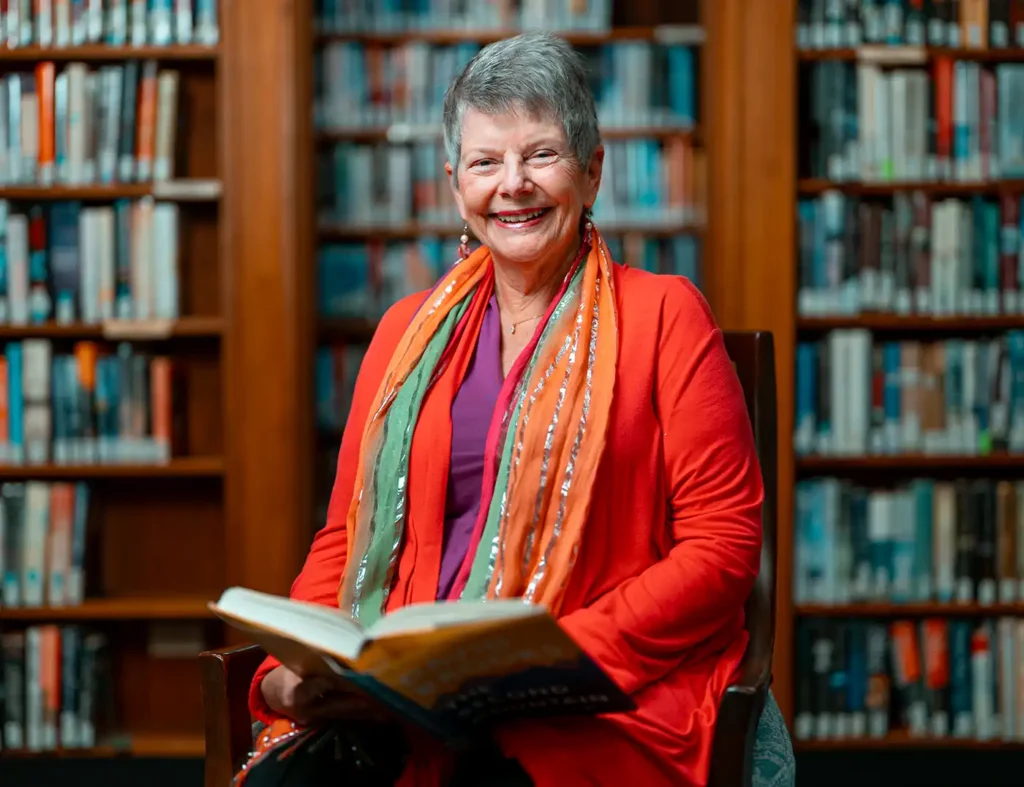 A senior woman poses in a library with a large book