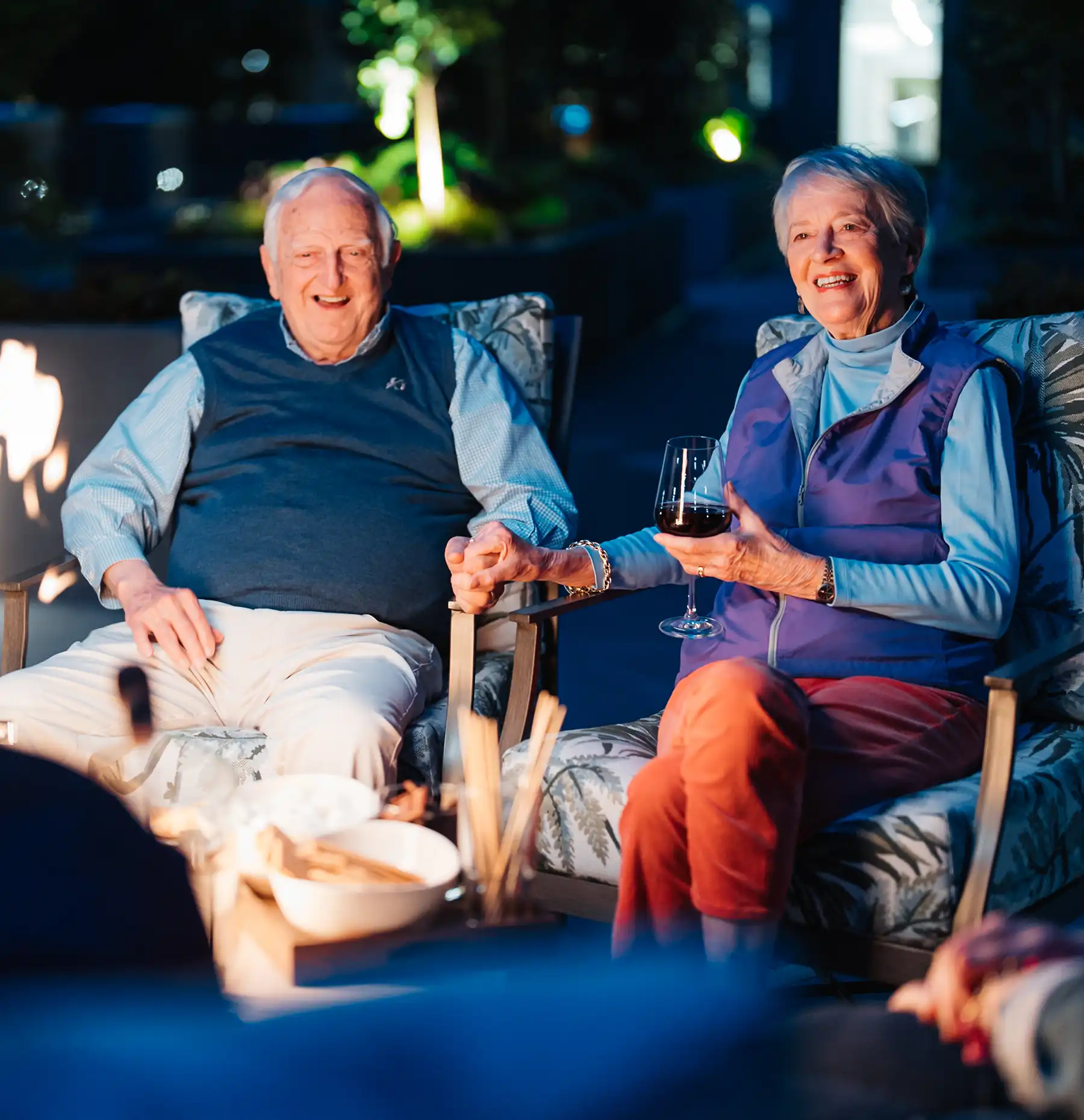 A senior couple hold hands seated by an outdoor fireplace